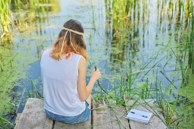 Mulher jovem sentada de costas no píer de madeira, apreciando a vista pitoresca da água do lago, relaxando na natureza em um dia ensolarado de verão, copie o espaço