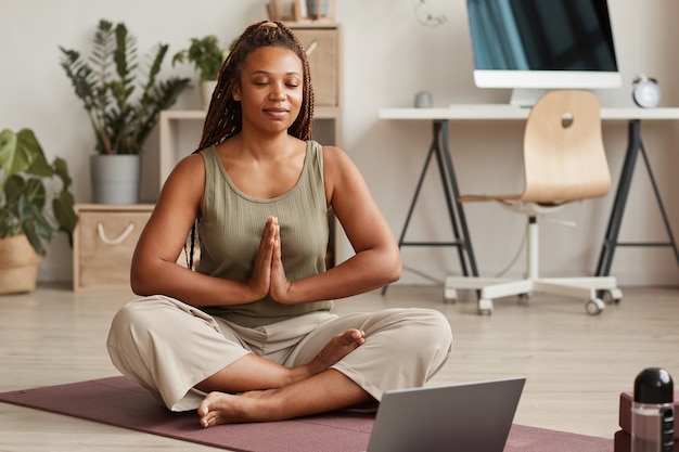 Mulher jovem sentada em posição de lótus na esteira de exercícios com os olhos fechados e meditando na sala de estar