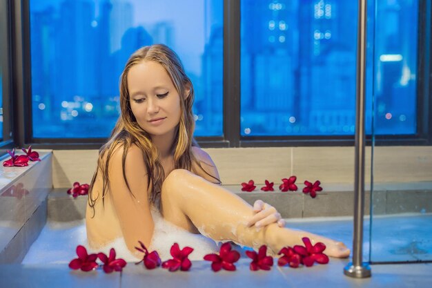 Mulher jovem sentada em uma banheira com flores de espuma e frangipani no fundo de uma janela panorâmica com vista para os arranha-céus e uma grande cidade à noite