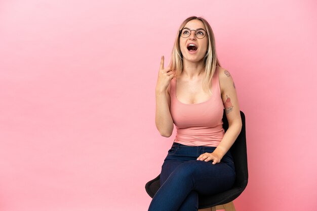 Mulher jovem sentada em uma cadeira sobre um fundo rosa isolado apontando para cima e surpresa