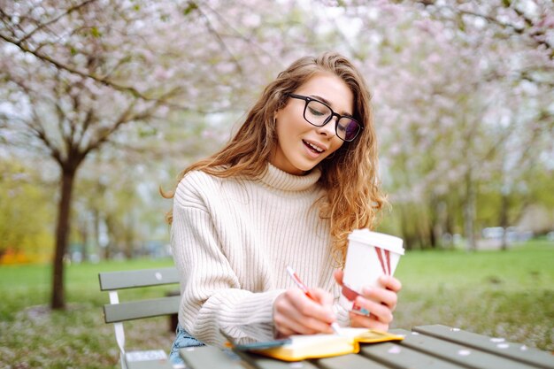 Mulher jovem sentada em uma mesa em um parque de primavera em flor com um bloco de notas e um laptop conceito de estilo de vida