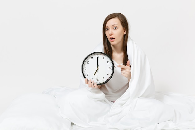 Mulher jovem sentada na cama com relógio redondo, lençol branco, travesseiro, embrulhado em cobertor na parede branca