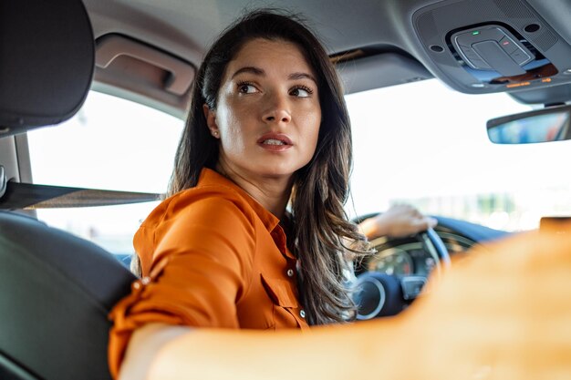 Mulher jovem sentada no assento do motorista no carro e olhando para a câmera por cima do ombro Retrato de mulher agradável com expressão positiva satisfeita com uma viagem inesquecível de carro