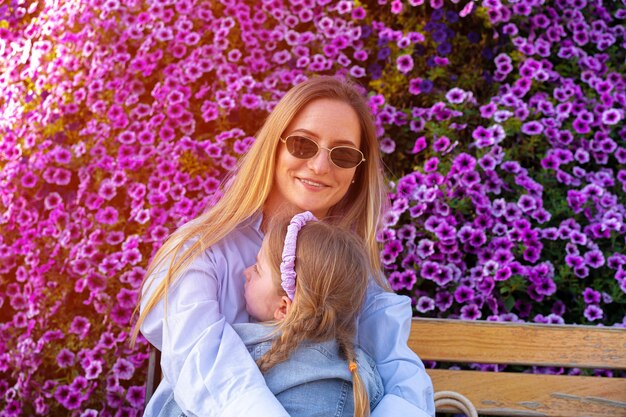 Mulher jovem sentada no banco com sua filha perto de uma parede de flores de petúnia