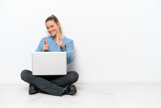 Mulher jovem sentada no chão com um laptop apontando para a frente e sorrindo