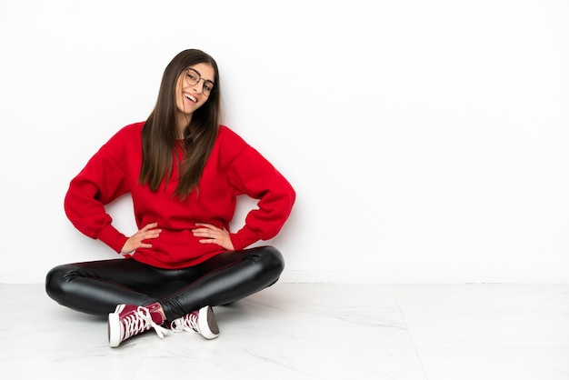 Mulher jovem sentada no chão, isolada no fundo branco, posando com os braços na cintura e sorrindo