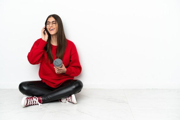 Mulher jovem sentada no chão, isolada no fundo branco, segurando um café para levar e um celular