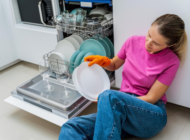 Foto mulher jovem sentada no chão perto da máquina de lavar pratos