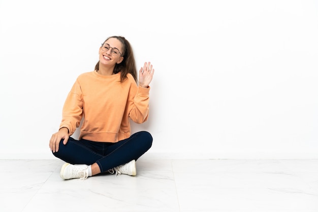 Mulher jovem sentada no chão saudando com a mão com expressão feliz