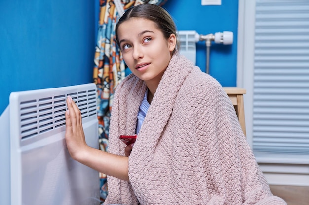 Mulher jovem sentada perto de um aquecedor elétrico em casa Menina adolescente se aquecendo sob um cobertor temporada de aquecimento crise de energia economizando frio outono conceito de temporada de inverno