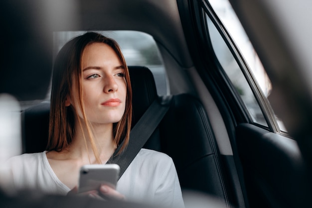 Mulher jovem séria com um smartphone sentado no carro e olhar para fora