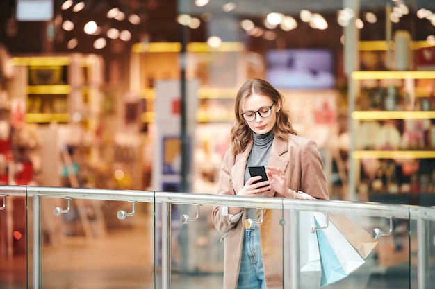 Mulher jovem séria de óculos em pé na grade e verificando o smartphone no shopping