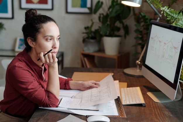 Mulher jovem séria de raça mista com coque de cabelo sentada na mesa e segurando a planta da casa enquanto analisa o esboço do projeto no computador