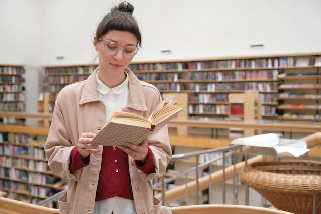 Mulher jovem séria examinando o livro em pé na grande biblioteca