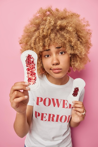 Mulher jovem séria segura dois tipos de absorventes higiênicos manchados com brilhos vermelhos como se o sangue falasse sobre menstruação e higiene feminina vestida com camiseta casual isolada sobre fundo rosa