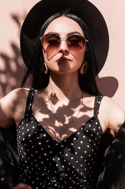 Mulher jovem sexy elegante com chapéu na jaqueta com vestido com óculos de sol gosta de sol na rua. Menina da moda do retrato da moda em preto elegante verão lindas roupas na luz do sol perto da parede rosa vintage.