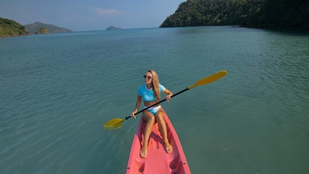 Mulher jovem sexy em maiô azul balança os pés na água na canoa rosa no mar contra a parte traseira das colinas