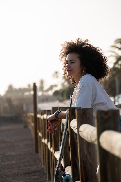 Mulher jovem skatista pensativa, apoiando-se em uma cerca ao pôr do sol