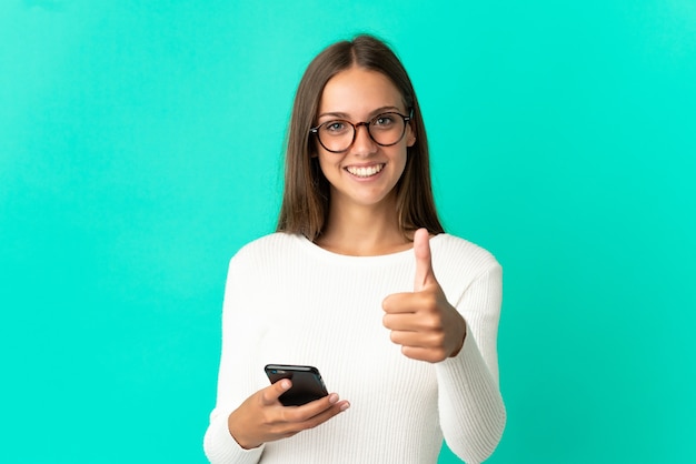 Mulher jovem sobre fundo azul isolado usando telefone celular enquanto faz o polegar para cima