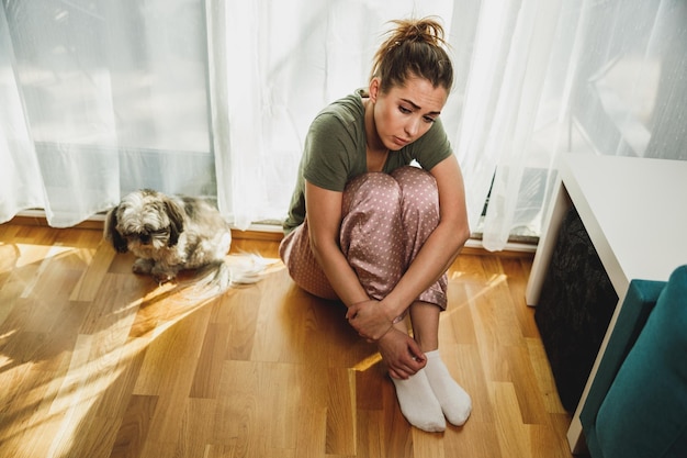 Mulher jovem solitária em depressão sente-se pela janela com os joelhos do abraço perdidos em maus pensamentos pessimistas.