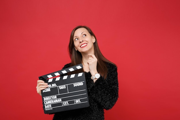 Mulher jovem sonhadora em suéter de pele preta, olhando para cima e segurando o clássico filme preto fazendo claquete isolada no fundo da parede vermelha. Emoções sinceras de pessoas, conceito de estilo de vida. Simule o espaço da cópia.