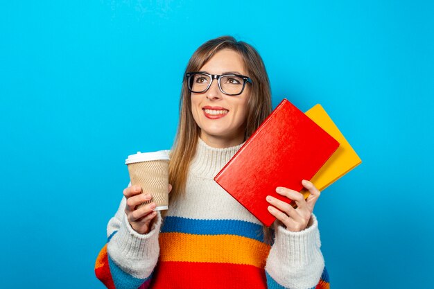 Mulher jovem sorri e segura livros e um copo de café ou chá nas mãos.