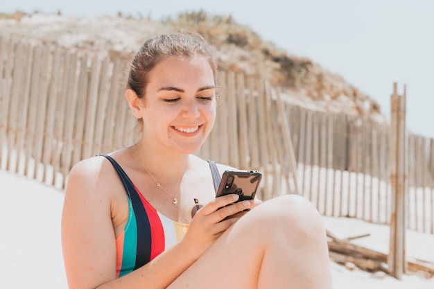 Mulher jovem sorri enquanto conversa ao telefone usando um maiô colorido na praia tomando banho de sol, conceito de viagens para jovens férias, espaço de cópia, rede social, conceito de usuário de óculos de sol