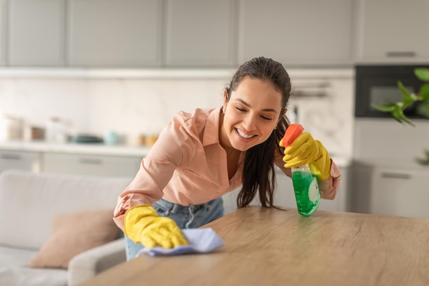 Mulher jovem sorridente a limpar a mesa da cozinha com spray