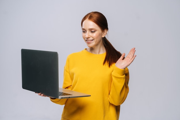 Mulher jovem sorridente, acenando com a mão e gesticulando olá para a tela do laptop, falando em videochamada com um amigo