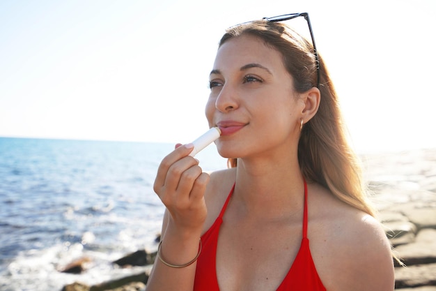 Mulher jovem sorridente, aplicando proteção solar nos lábios na praia