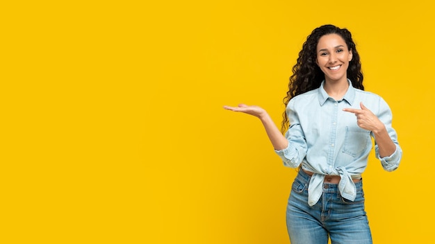 Mulher jovem sorridente apontando para o espaço em branco posando e sorrindo sobre o fundo amarelo do estúdio