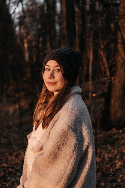 Mulher jovem sorridente com casaco e chapéu em pé na floresta de outono