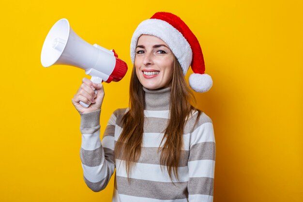 Mulher jovem sorridente com chapéu de Papai Noel com um megafone em um fundo amarelo.