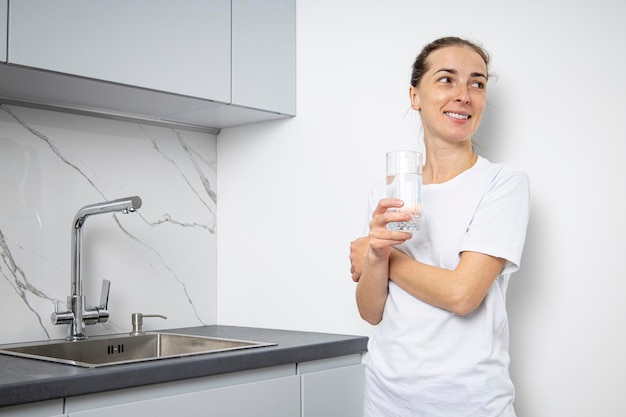 Mulher jovem sorridente com copo de água em pé na cozinha