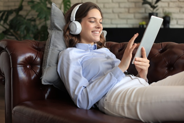 Mulher jovem sorridente com fones de ouvido e tablet digital no sofá.