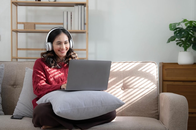 Mulher jovem sorridente com fones de ouvido usando laptop na sala de estar Mulher estudando em casa Jovem mulher usando fones de ouvido ouve curso de áudio online grátis em casa