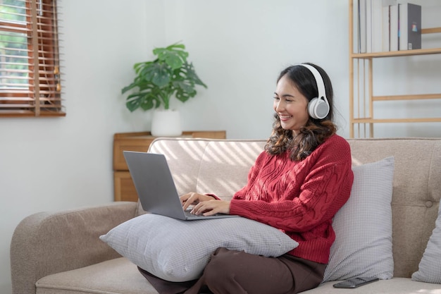 Mulher jovem sorridente com fones de ouvido usando laptop na sala de estar Mulher estudando em casa Jovem mulher usando fones de ouvido ouve curso de áudio online grátis em casa