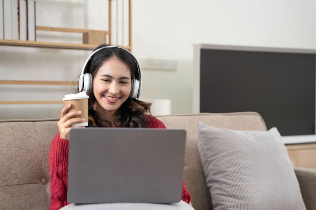 Mulher jovem sorridente com fones de ouvido usando laptop na sala de estar Mulher estudando em casa Jovem mulher usando fones de ouvido ouve curso de áudio online grátis em casa