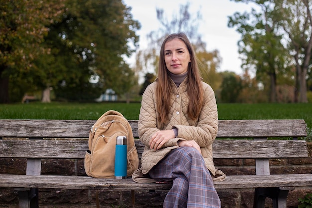 Mulher jovem sorridente com garrafa térmica sentado no banco do parque