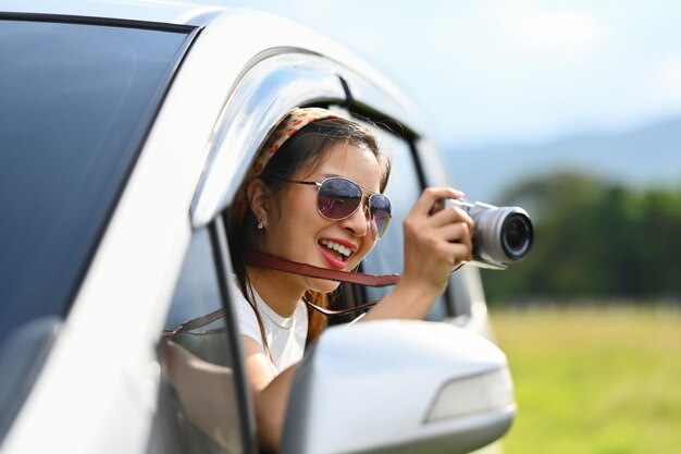 Foto mulher jovem sorridente com óculos de sol tirando foto com a câmera enquanto se inclinava na janela do carro durante