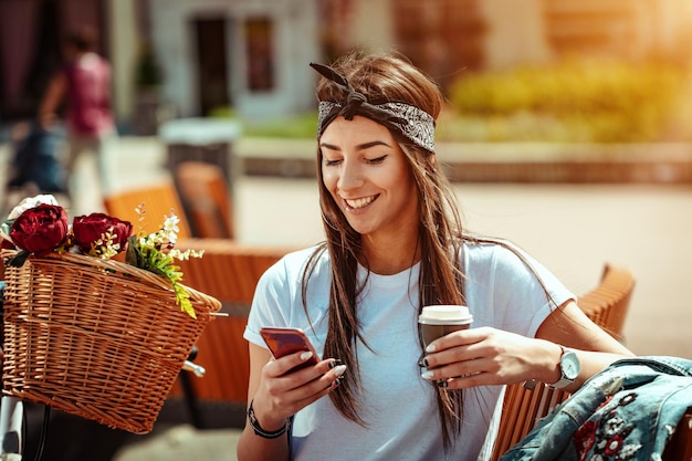 Mulher jovem sorridente com smartphone, tomando café e se senta em um banco em um dia ensolarado, ao lado da bicicleta com cesta de flores.