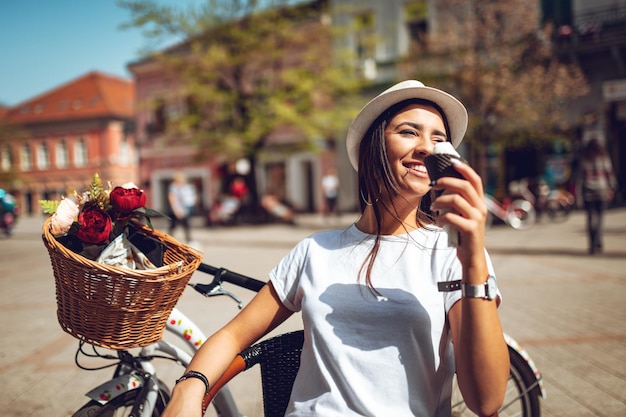 Mulher jovem sorridente com sorvete, fazendo uma pausa em uma praça da cidade em um dia ensolarado, ao lado da bicicleta com cesta de flores.