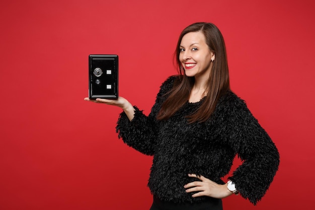 Mulher jovem sorridente com suéter de pele preta segurando o banco de metal seguro para o acúmulo de dinheiro isolado no fundo da parede vermelha brilhante no estúdio. Conceito de estilo de vida de emoções sinceras de pessoas. Simule o espaço da cópia.