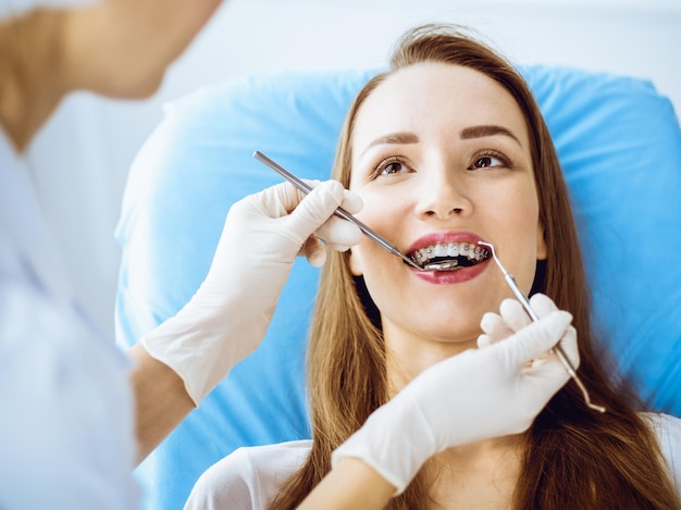 Mulher jovem sorridente com suportes ortodônticos examinados pelo dentista na clínica odontológica. Dentes saudáveis e conceito de cuidados médicos.