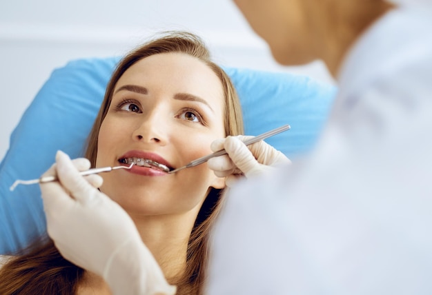 Mulher jovem sorridente com suportes ortodônticos examinados pelo dentista na clínica odontológica. Dentes saudáveis e conceito de medicina.