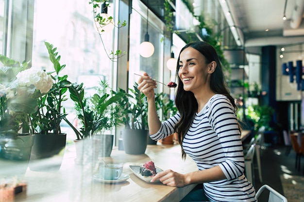 Mulher jovem sorridente comendo bolo de chocolate com sorvete de frutas e café no café