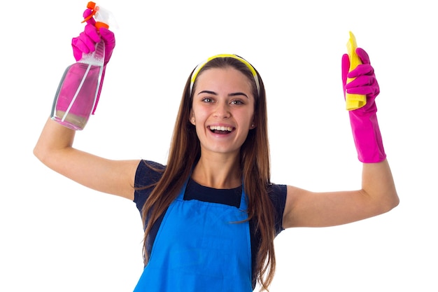 Foto mulher jovem sorridente de camiseta azul e avental com luvas cor de rosa segurando espanador amarelo e detergente em fundo branco no estúdio