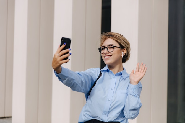 Mulher jovem sorridente de óculos e camisa azul, usando o smartphone para uma conversa com vídeo no ar fresco. Conceito de comunicação e tecnologia à distância.