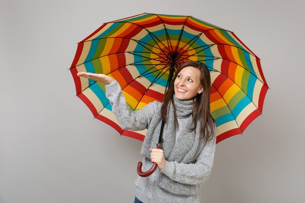 Mulher jovem sorridente de suéter cinza, lenço apontando a mão segurando guarda-chuva colorido isolado no fundo da parede cinza. emoções de pessoas de estilo de vida de moda saudável, conceito de estação fria. simule o espaço da cópia.