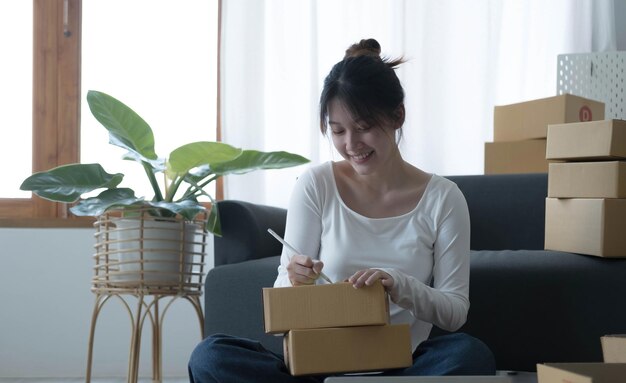 Mulher jovem sorridente dona de negócios asiáticos prepara a caixa de encomendas e verifica os pedidos on-line de produtos para entregar ao cliente no laptop Conceito de compras on-line